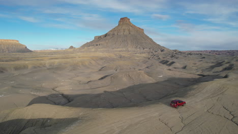 Toma-De-Drone-De-Un-Vehículo-Todoterreno-Rojo-Moviéndose-Sobre-Una-Colina-En-El-Desierto-De-Utah-Con-Una-Formación-Rocosa-De-Fábrica-En-El-Fondo