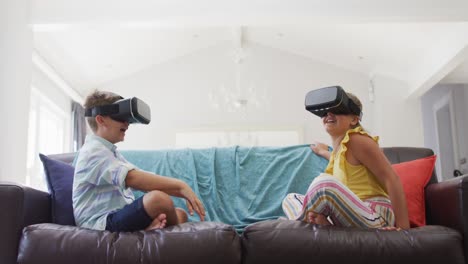 happy caucasian brother and sister at home, sitting on couch using vr headsets and smiling