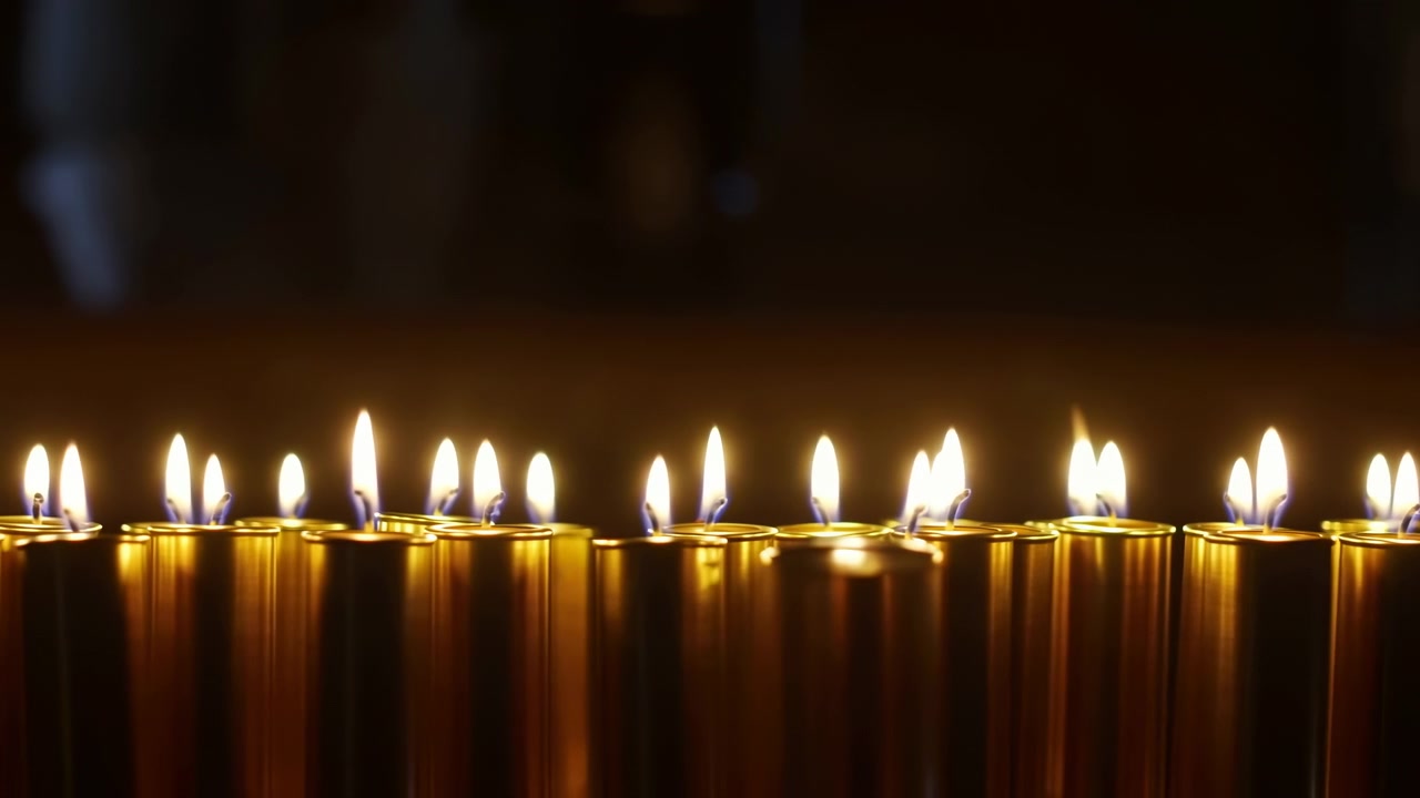Scena ravvicinata in movimento di più candele tealight accese disposte in fila orizzontale su uno sfondo scuro, con sottili variazioni nell'altezza e nell'intensità della fiamma e morbidi riflessi su coppe dorate per la calma