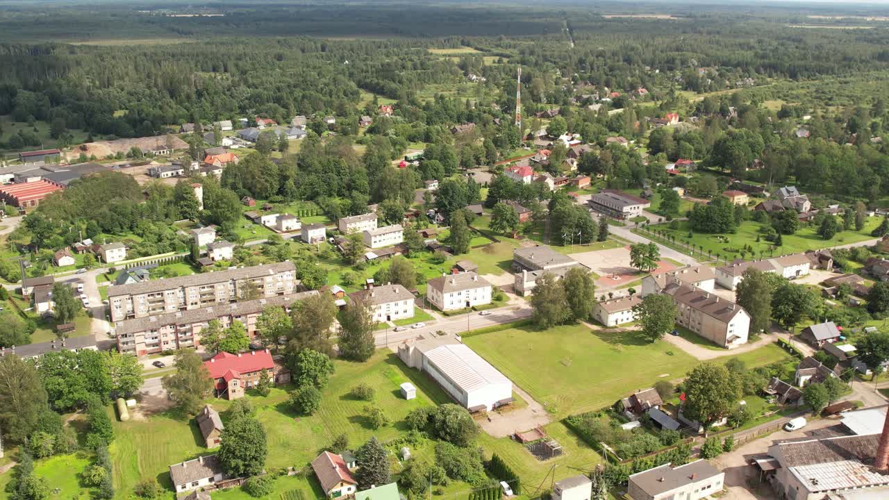 Aerial view of a small town hosting a lively outdoor fair with market stalls, parked cars, houses, and surrounding green fields under a sunny sky. Myisakula
