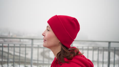 side view of athlete jogging in red beanie and jacket along snowy pathway near iron railing, overlooking urban cityscape in foggy atmosphere, with bridge and cars in background