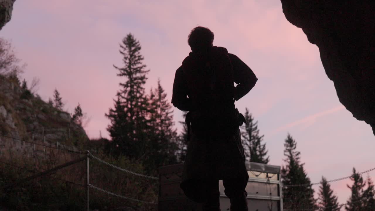 Drammatica silhouette di un escursionista di montagna che emerge dall'apertura di una grotta rocciosa durante il tramonto all'ora d'oro nelle Alpi. Il cielo rosa-violaceo crea una splendida retroilluminazione attraverso un arco di pietra naturale con alberi sempreverdi.