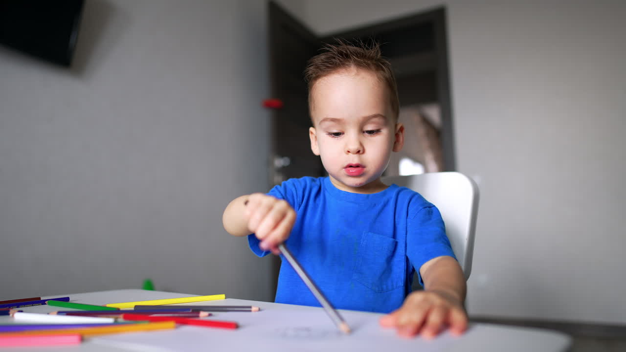 Charming baby boy sitting at desk holding a pencil. Cute kid drawing and talking. Blurred backdrop.