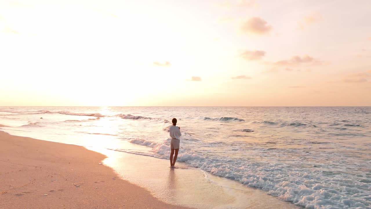 A stunning bird’s-eye perspective captures a lone woman standing barefoot on the white sands of Geiymiskih Fannu Beach as the Maldivian sun dips below the horizon.