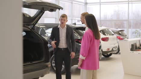 young car salesman showing to young couple new automobile at dealership salon.