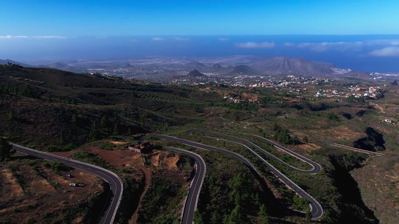 Vista aerea di una strada di montagna tortuosa attraverso terreno vulcanico con una città costiera in lontananza.