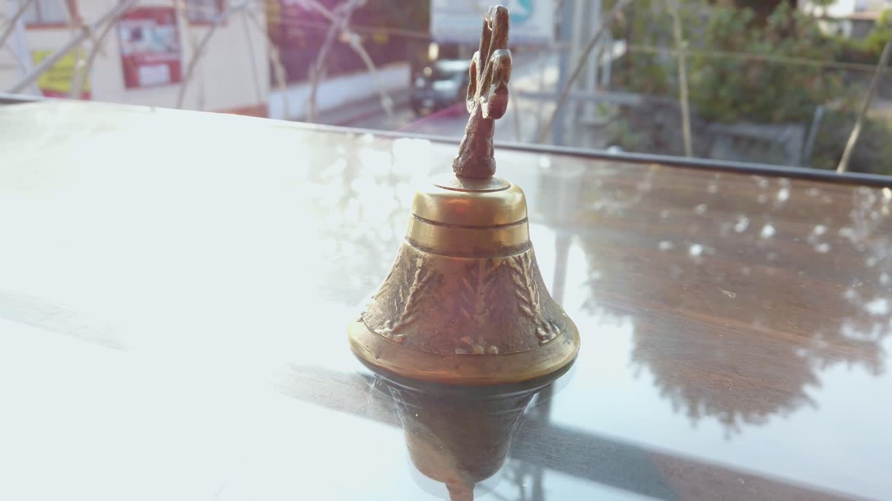 Inquadratura semi-orbitale attorno a un'antica campana di ottone con manico a forma di trifoglio, appoggiata su una superficie riflettente all'aperto.