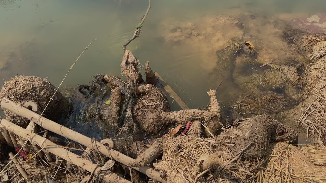 Mud-covered straw remains of a submerged idol rest along a calm riverbank, captured in a static shot showing post-immersion debris and textures