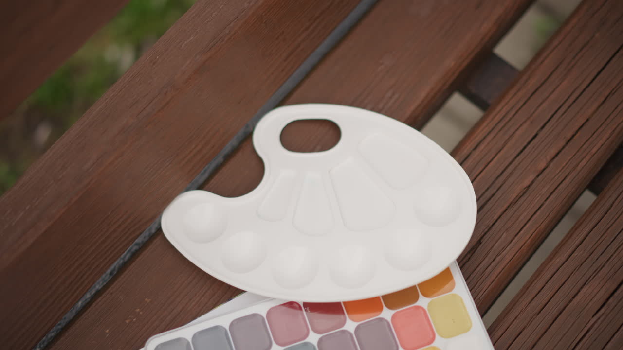 Set di acquerelli con tavolozza bianca su una panca, un artista posiziona una tavolozza di plastica vuota accanto a un foglio di campioni pastello su una panca di legno a doghe, luce solare naturale e soffusa, tonalità di colore tenui, un tranquillo momento creativo.