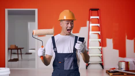 Asian Man Painter Wearing Safety Helmet Looking At Smartphone, Smiling, And Saying Wow While Standing With Painting Wall In Room