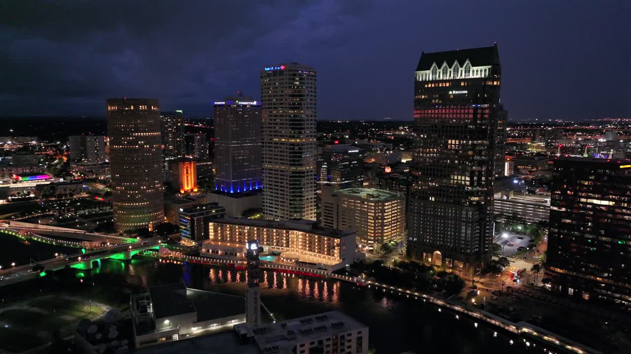 Tampa’s skyscrapers along the Hillsborough River at night. Lights reflect on the water. Prominent buildings and bridge in view. Aerial perspective.