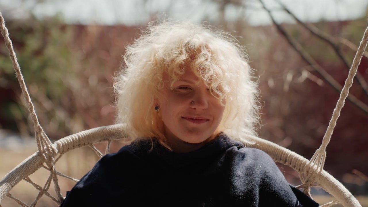 Pessoa jovem com cabelo loiro encaracolado sentada em cadeira de vime suspensa ao ar livre, girando suavemente enquanto a brisa levanta os cachos, cordas permanecem esticadas, e a luz quente cria um clima calmo e relaxado em meio a um jardim natural