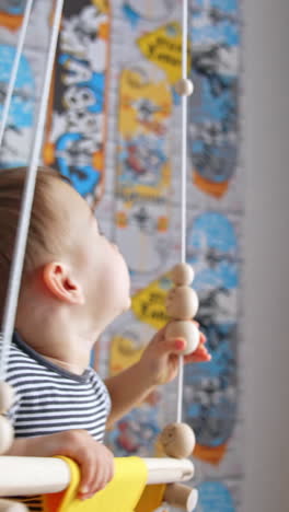 little boy swings in a yellow swing indoors. kid plays with wooden beads on the rope. vertical video