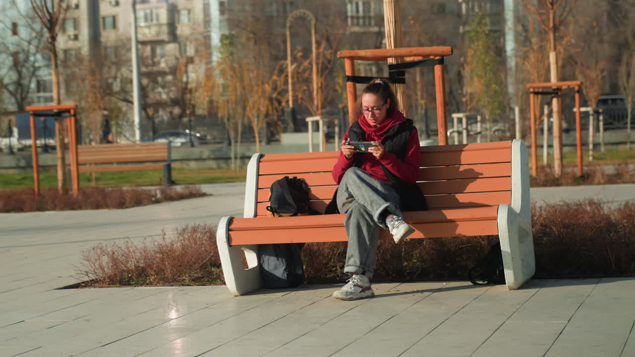 Žena na lavičce čte telefon v parku, studentka ponořená do zprávy, červená bunda, batoh vedle, měkké podzimní světlo, prázdné náměstí a betonová cesta, spadané listí a zamyšlená nálada.