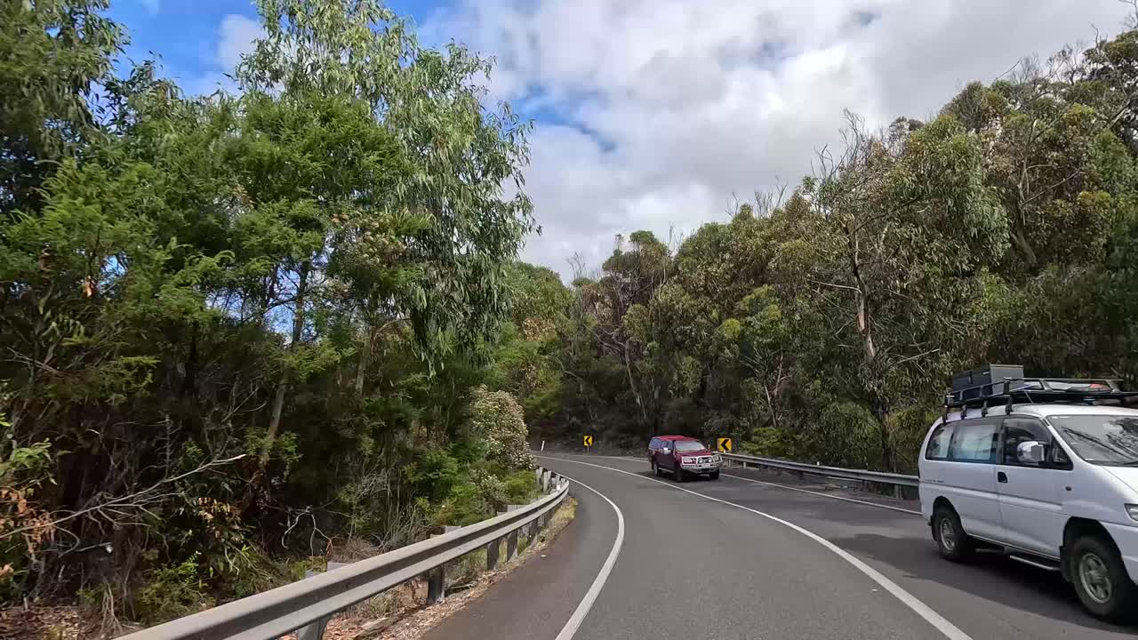 Vehicles travel through lush greenery and winding roads under a bright sky, capturing the serene beauty of the Great Ocean Road