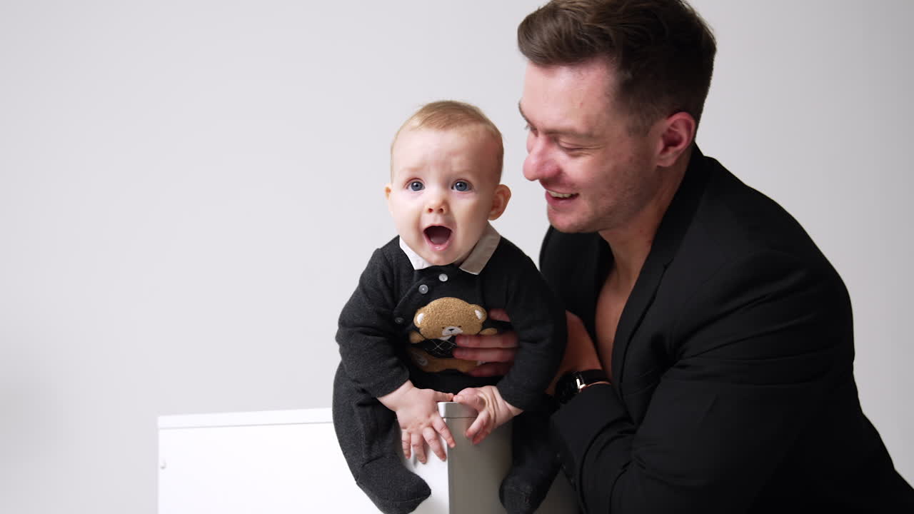 Adorable little baby sits on the cube and opens his mouth wide. Father holding his kid and kisses him on the head. White backdrop.