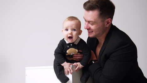 adorable little baby sits on the cube and opens his mouth wide. father holding his kid and kisses him on the head. white backdrop.