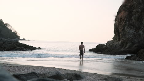 man slowly walks into ocean in small peaceful cove at sunset, slow motion