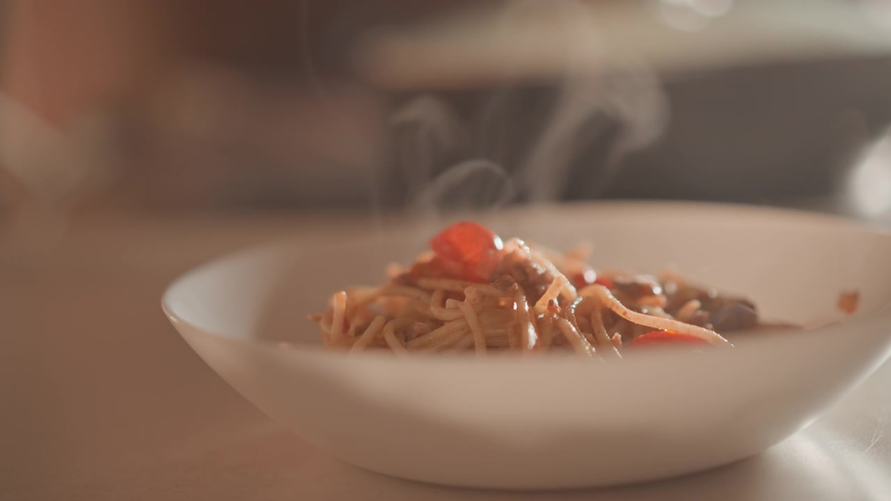 Primo piano di spaghetti caldi in ciotola bianca, con salsa lucida che avvolge la pasta mentre un leggero vapore sale, guarniti con pomodorini e erbe aromatiche che aggiungono colore, sotto una luce calda e appetitosa.