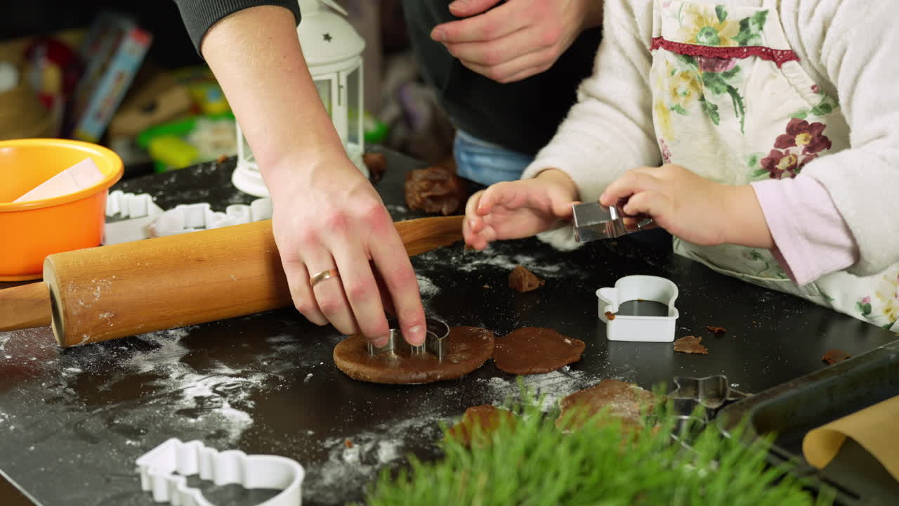 Festive family moment showing mother helping young child cutting gingerbread shapes with cookie cutters, flour dusted wooden surface and holiday decor surrounding baking scene