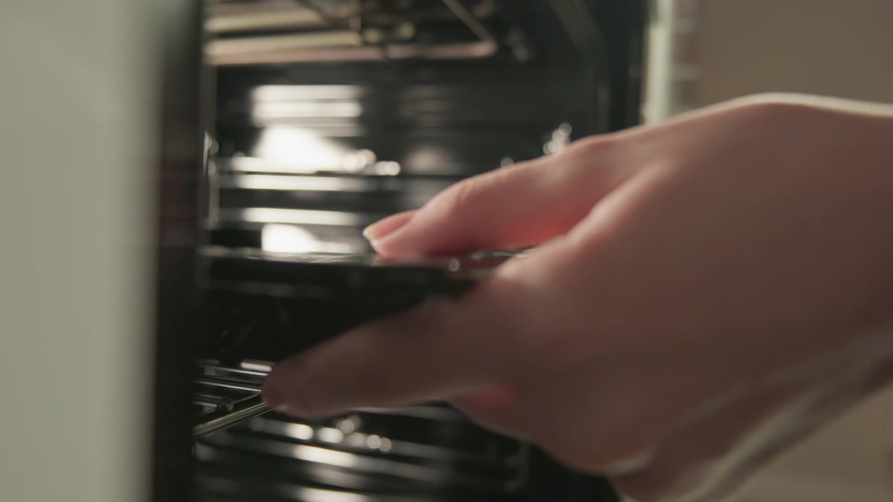 Una vista ravvicinata mostra le mani che fanno scivolare una teglia nella cavità di un forno aperto, con luce interna calda, griglie metalliche, superfici lucide e un movimento in avanti visibile durante un'attenta preparazione in cucina.