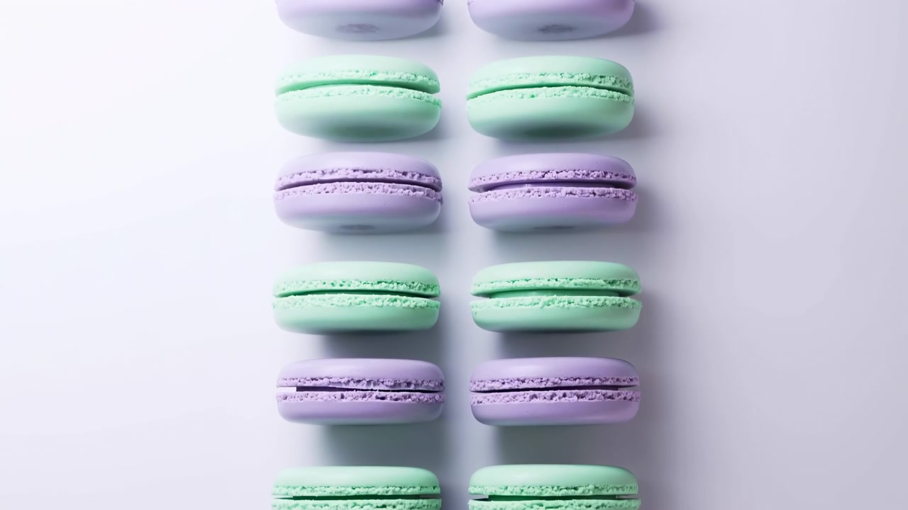 Two vertical columns of intact pastel macarons alternating mint green and lavender lie horizontally on near-white studio surface, showing cream filling between smooth domed shells and textured