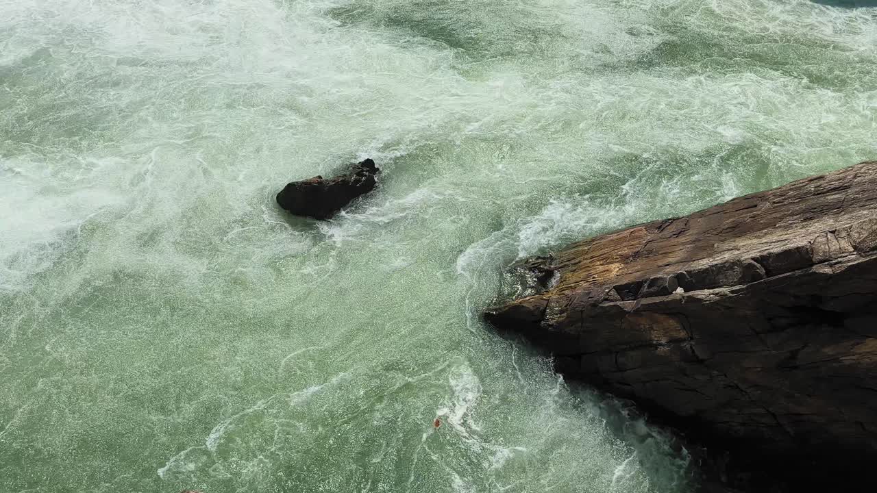 Topbillede af hurtigtstrømmende flodvand, der hvirvler omkring mørke sten, viser turbulente strømme, skummende teksturer og kraftig naturlig strøm i et stenet flodløb