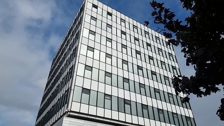Moderno edificio per uffici universitari nel centro con facciata in vetro che riflette il cielo blu nuvoloso, Lenta inclinazione verso il basso