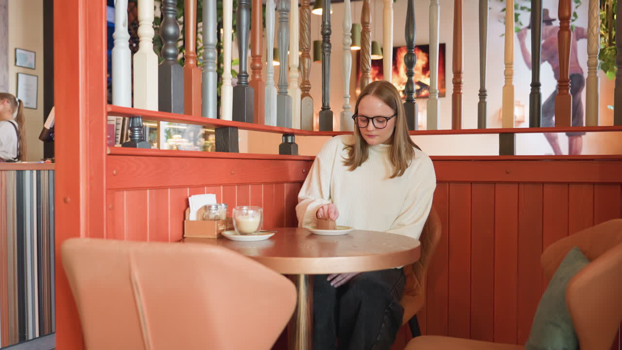 young woman in white sweater sits at mall cafe table behind decorative railing, eats chocolate cake while sipping latte, modern mall bar decor