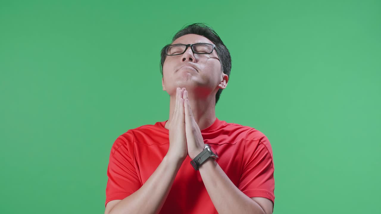 눈을 닫은 으로 쳐다보는 모습: 공을 들고 있는 아시아인 남자가 팀을 위해 기도하다가 손을 들고 초록색 스크린 배경에서 축구 경기를 응원하고 있다.