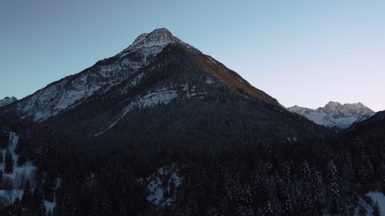 Flying away from huge and impressive mountain with forest and trees in winter with snow and ice in the idyllic and scenic Bavarian Austrian alps mountains with red sunset peaks