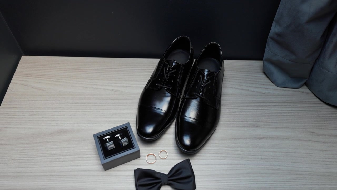 Smooth camera pans over sleek black shoes, rings, bow tie and cufflinks on a wood dresser. Stylish wedding accessories laid out with soft, even lighting for modern detail shots. (199)