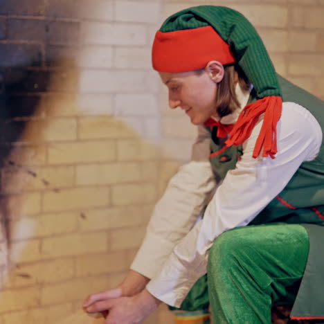 man in a festive costume of elf is putting logs into the open fire in fireplace. bright fire burning in fireplace and a young elf is sitting near it on a big log in the house. christmas time.