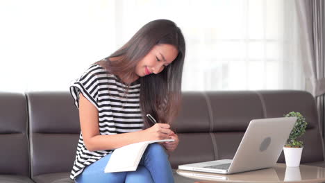 asian thai woman doing taxes at home with a laptop and write down notes in a notebook