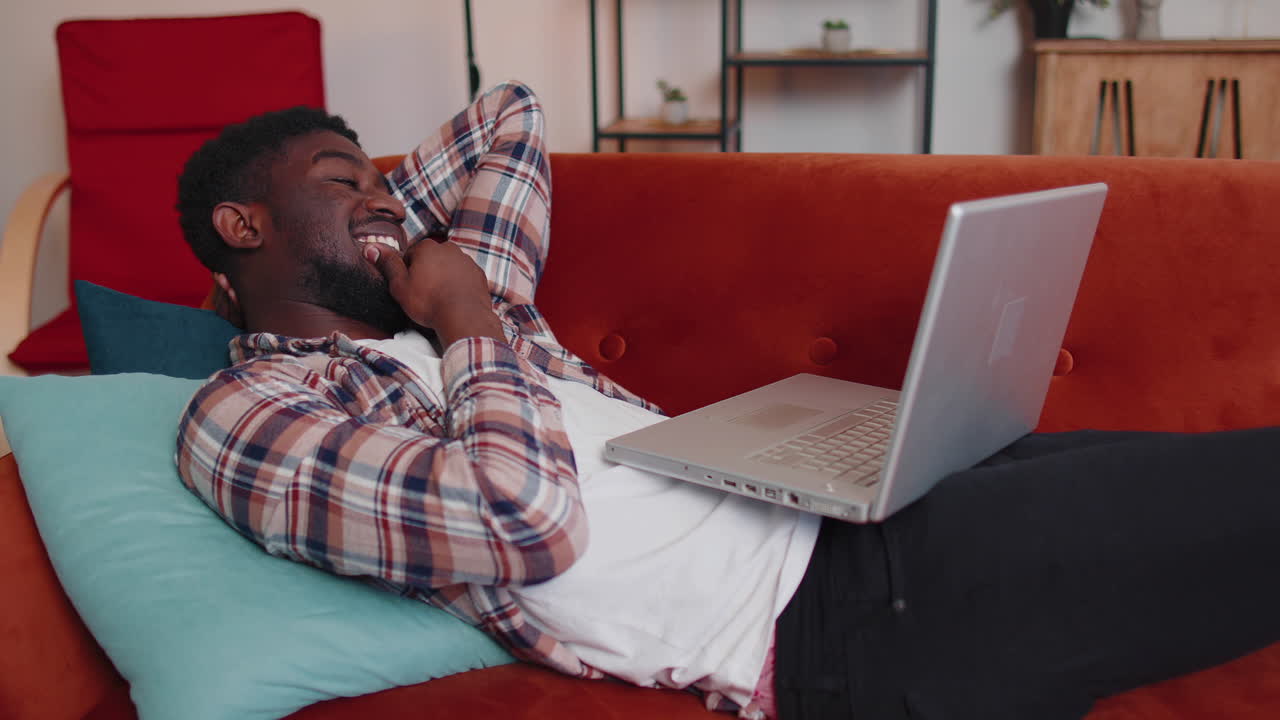 Young man using laptop computer lying on sofa at home working, online shopping, watching movies