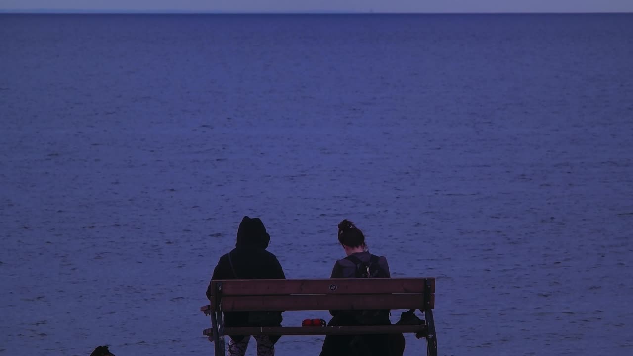 Two people and a dog sitting on a bench by the water at dusk