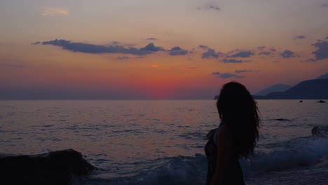 Silueta-De-Mujer-Hermosa-Con-El-Pelo-Rizado-De-Pie-En-La-Playa-Rocosa,-Viendo-El-Colorido-Atardecer