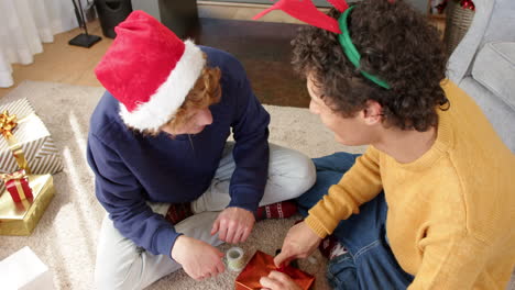 happy diverse gay male couple wrapping christmas presents by fireplace at home, slow motion