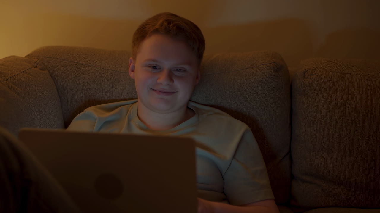 Teen White Boy On Couch Smiling At Laptop Screen, Evening Warm Lamp Glow, Relaxed Posture In Casual Tshirt,