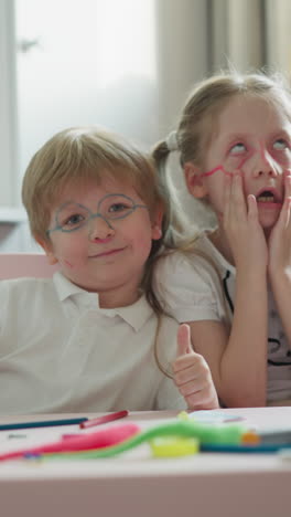 couple of children with painted faces sits at desk in classroom. boy shows thumbs-up smiling cutely. girl grimaces and rolls eyes making funny faces