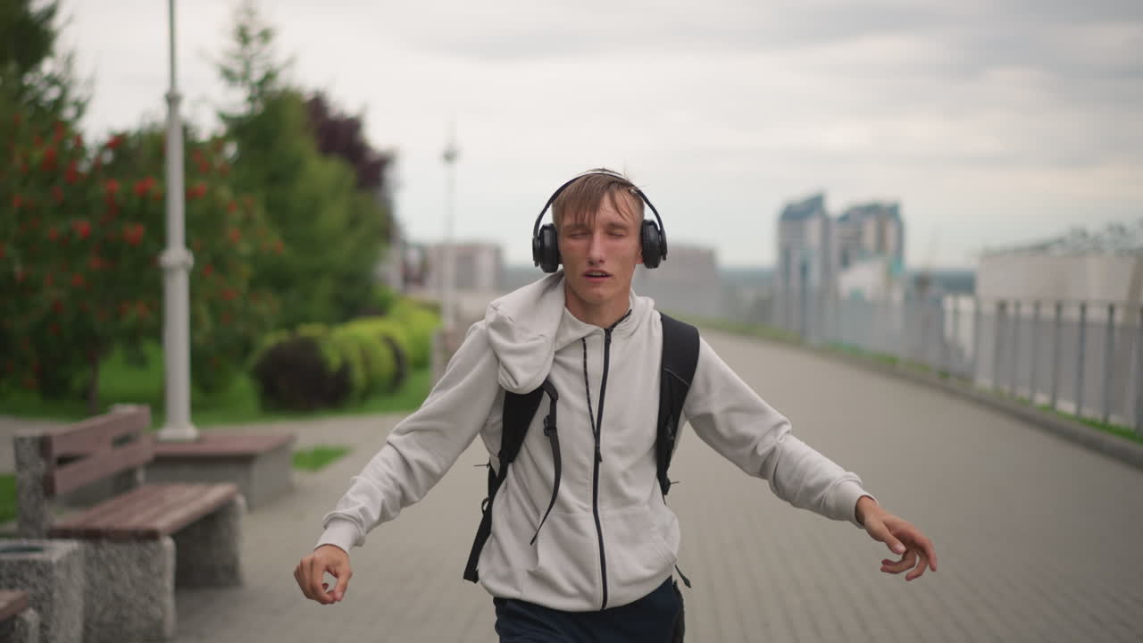 homem branco conferindo as horas no relógio, calçadão nublado, fones de ouvido, vista frontal, expressão mudando para sorriso, ajusta o capuz e o headset, caminha em direção à câmera, energia otimista, aproximando-se do horizonte urbano, retrato espontâneo