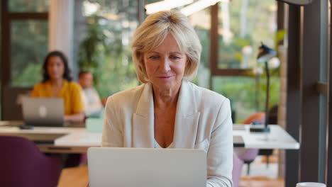 mature businesswoman working on laptop at desk in office pausing to look out of window