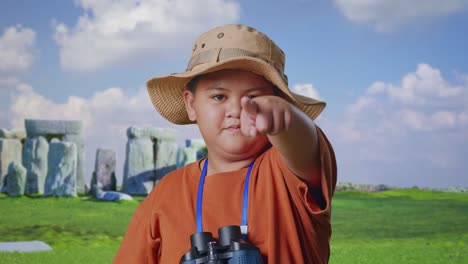 asian boy with hat and binoculars using magnifying glass pointing finger towards you, shaking head waving index finger while traveling in stonehenge. boy researcher showing no gesture, close up