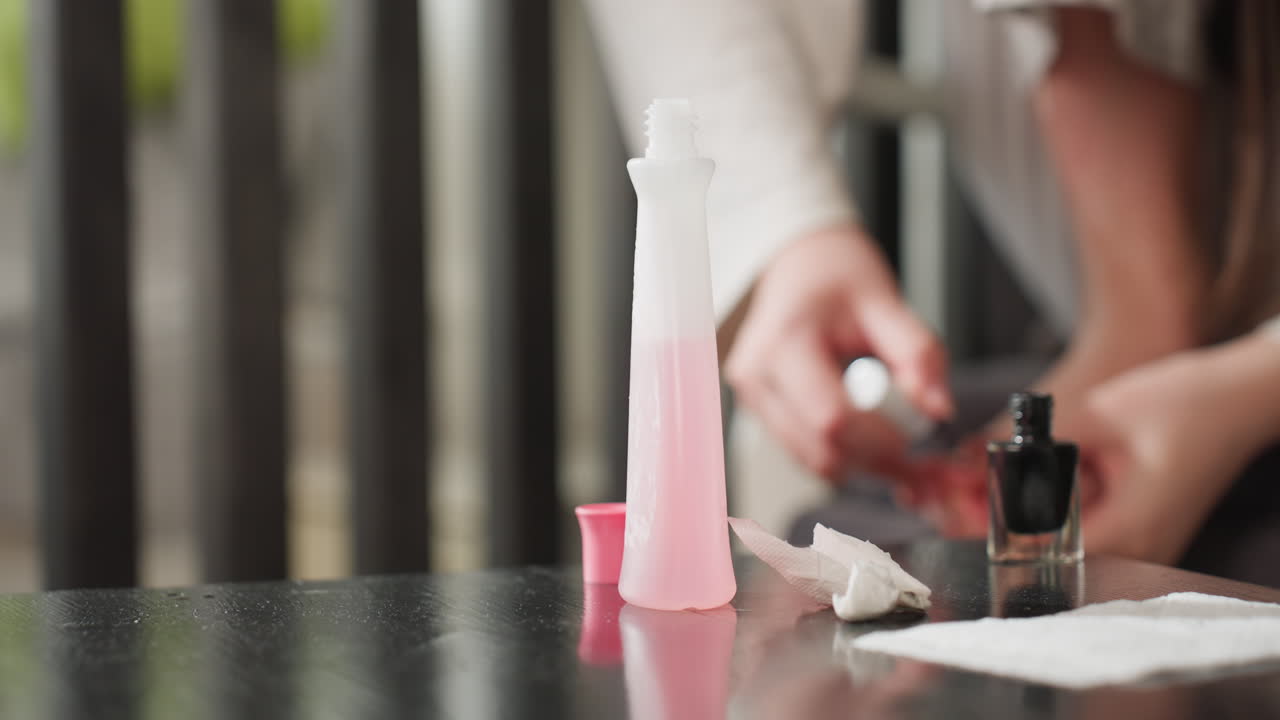 Close view shows girl on chair rubbing black polish on toes, remover bottle with open cap and tissue on glossy table, stained wipe nearby, soft daylight, careful pedicure, steady hands