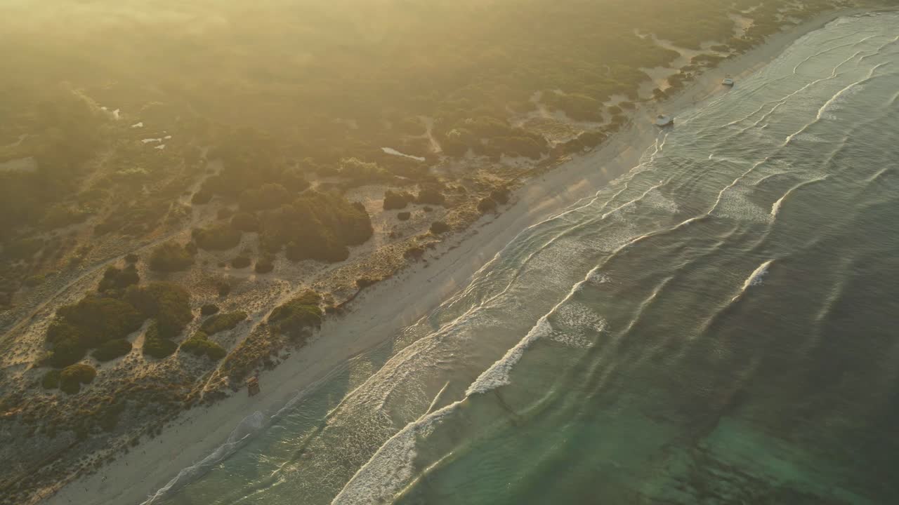 A golden sunset bathes a pristine beach in warm light, revealing gentle waves lapping against the shore and dense vegetation covering nearby sand dunes.