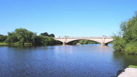 Vehicles-travelling-over-an-arched-stone-bridge-with-a-blue-lake-below