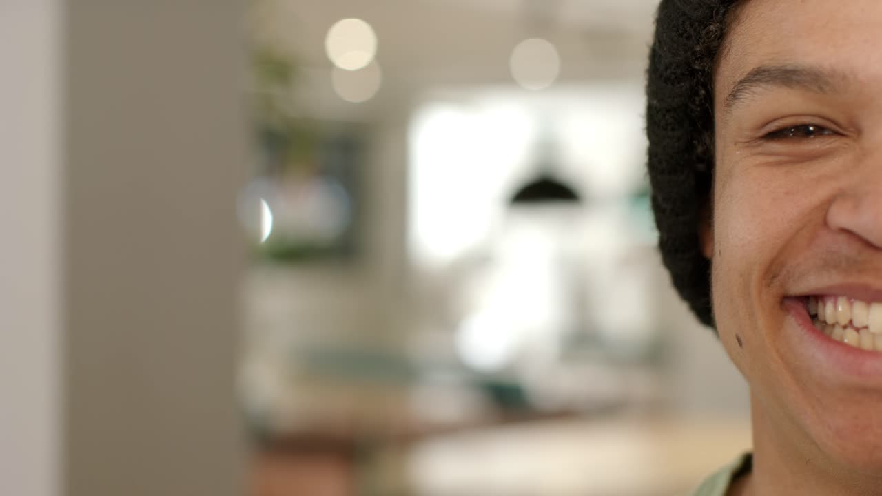 Man is hearing offscreen joke and is turning smile into laugh while wearing beanie near window
