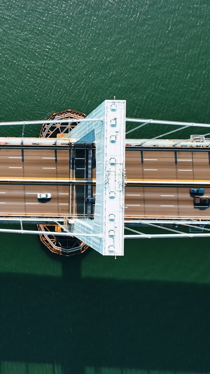 Descending over the powerful support of a suspension bridge. View of six-lane road with multiple cars driving by. Top perspective. Vertical video