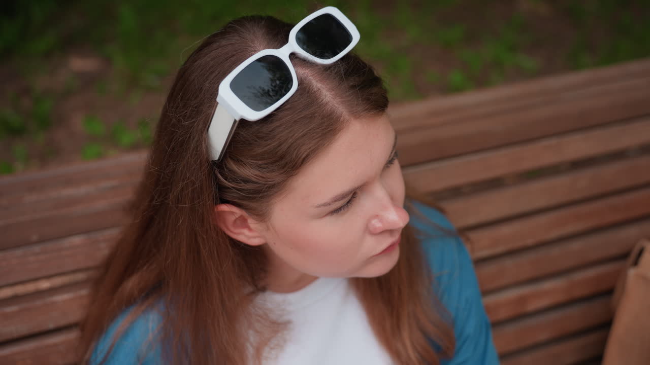 Una mujer disfruta de un plátano sentada en un banco de madera en un parque soleado, con un ambiente relajado bajo los árboles verdes, luz natural que resalta los tonos de la cara y el pelo, con un atuendo informal que refleja un estilo de vida veraniego tranquilo.