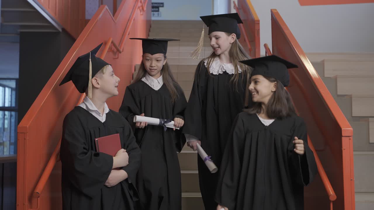 모자와 가운을 입은 행복한 백인 미취학 남학생이 책을 들고 카메라를 쳐다보는 동안 그의 반 친구들이 졸업식에서 계단을 내려가는 동안